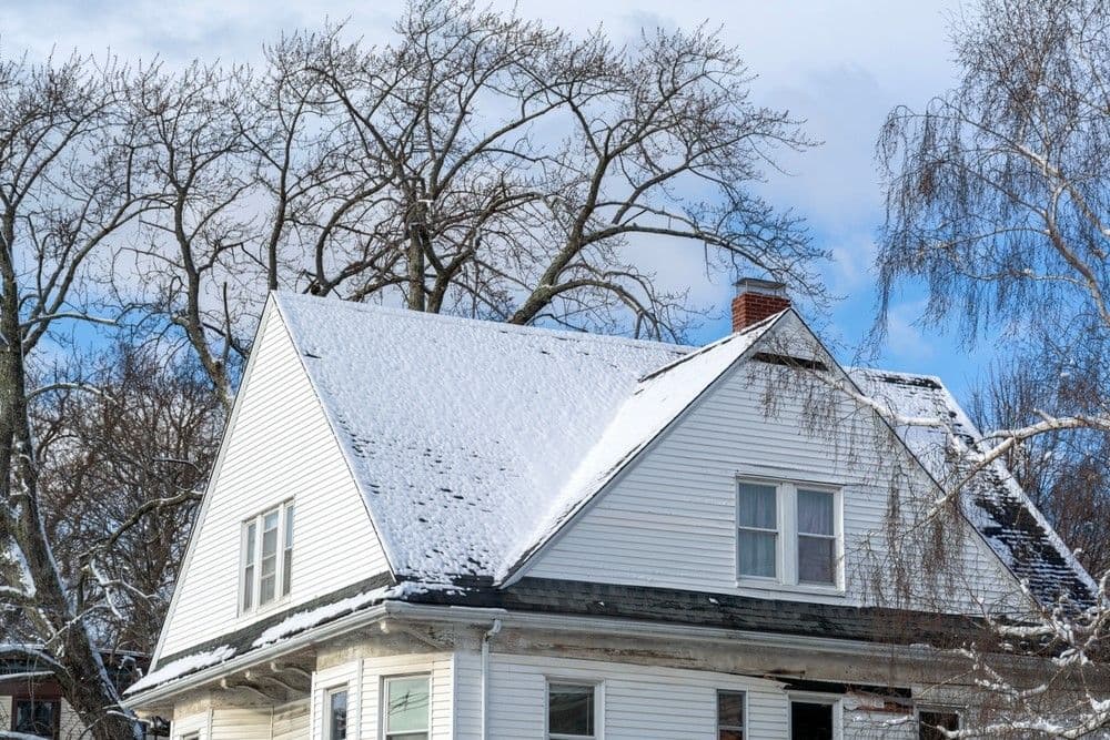 A quaint, white, two-story house with a snow-covered roof and dormer windows set against a backdrop of bare, winter trees and a blue sky.