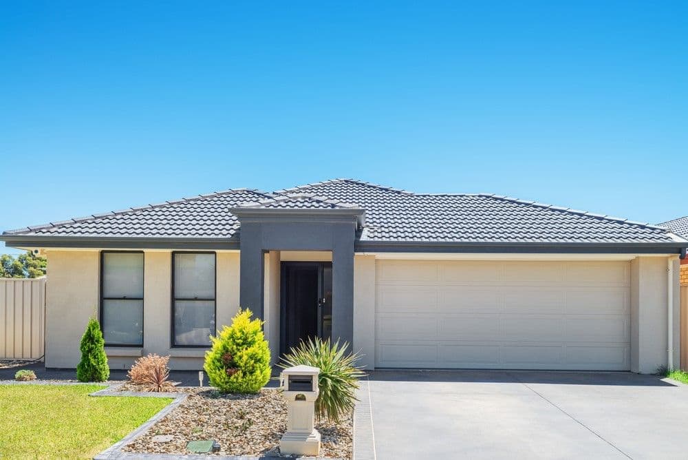 Modern single-story house with a gray tiled roof, beige walls, and a large garage. Neatly landscaped front yard with shrubs under a clear blue sky.