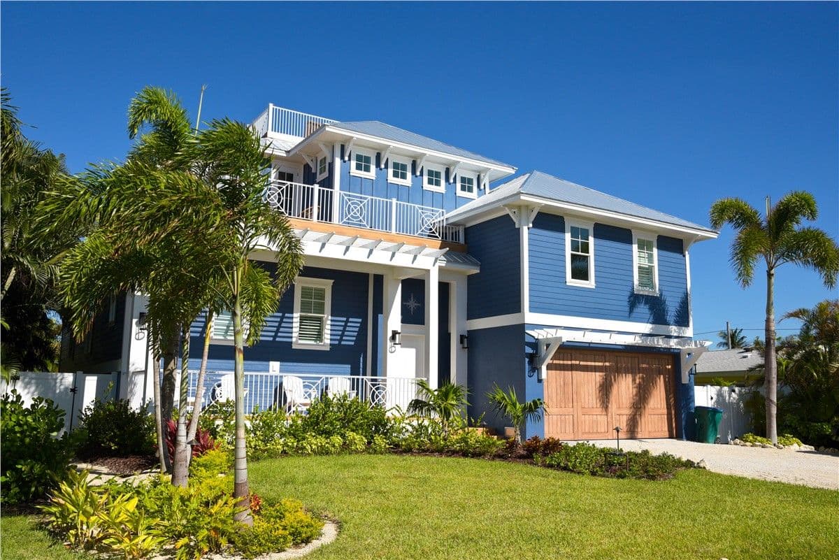 Two-story blue house with white trim, surrounded by lush palm trees and a well-maintained garden. A bright, sunny day enhances the welcoming atmosphere.