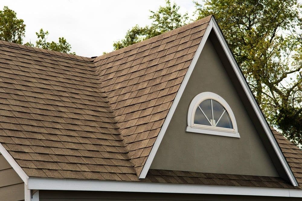 A brown shingled roof with an arched window at the peak, set against a backdrop of green trees and a cloudy sky, conveys a serene suburban atmosphere.