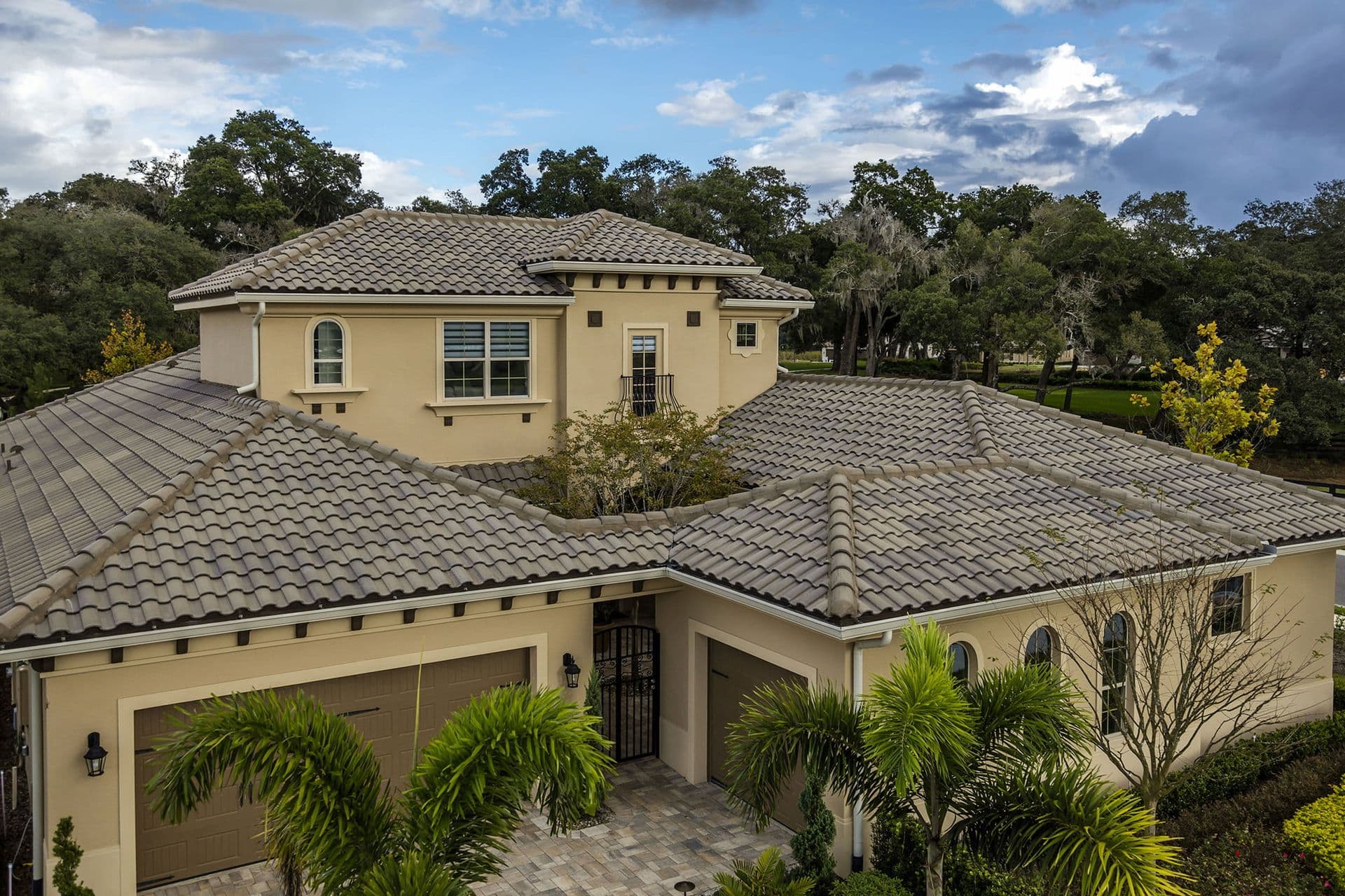 Aerial view of a two-story beige house with a gray tiled roof, surrounded by lush trees and palm plants. The sky is partly cloudy, creating a serene atmosphere.