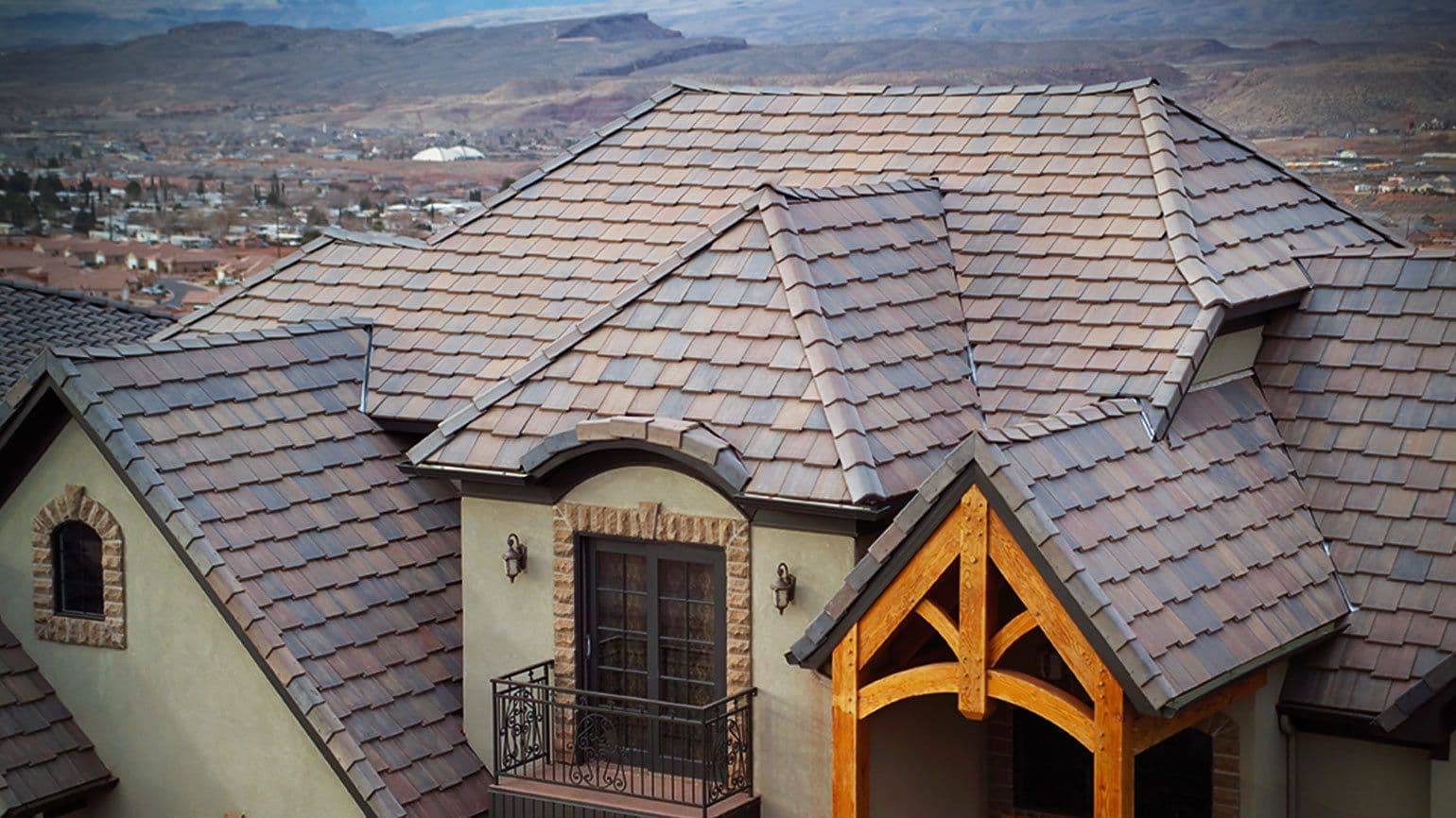Aerial view of a house with a complex, multi-angled roof made of brown shingles. The background shows a sprawling town and distant mountains.