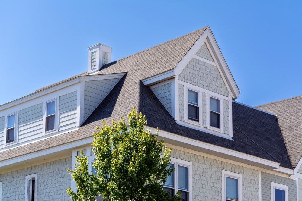 Gray two-story house with white trim, featuring a steep gabled roof and several windows. A leafy tree partially obscures the facade. Clear blue sky.