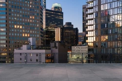 View of a cityscape at dusk featuring modern skyscrapers with illuminated windows. A concrete terrace in the foreground adds depth to the urban scene.