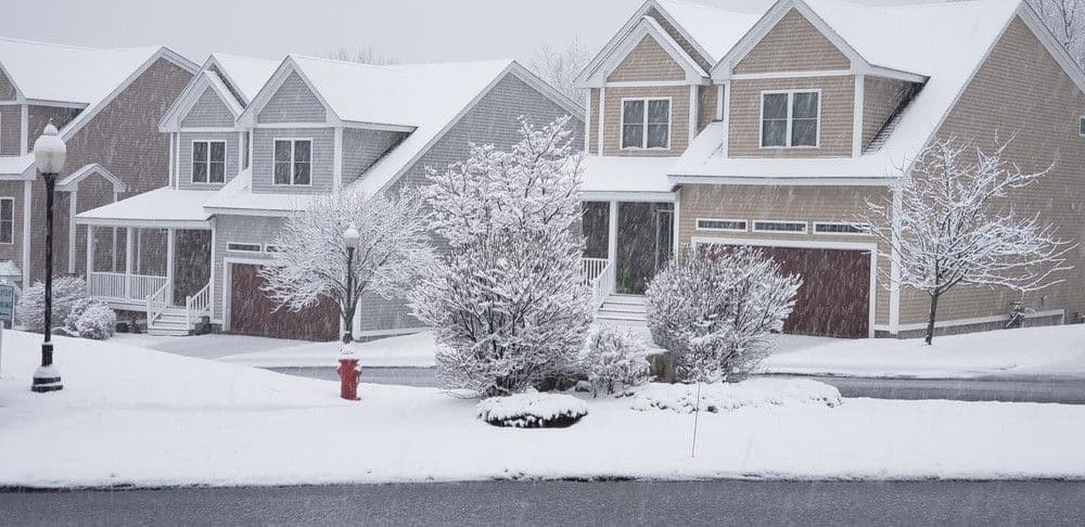 Snow-covered suburban street with two-story houses, bare trees, and a fire hydrant. The scene conveys a quiet, serene winter atmosphere.