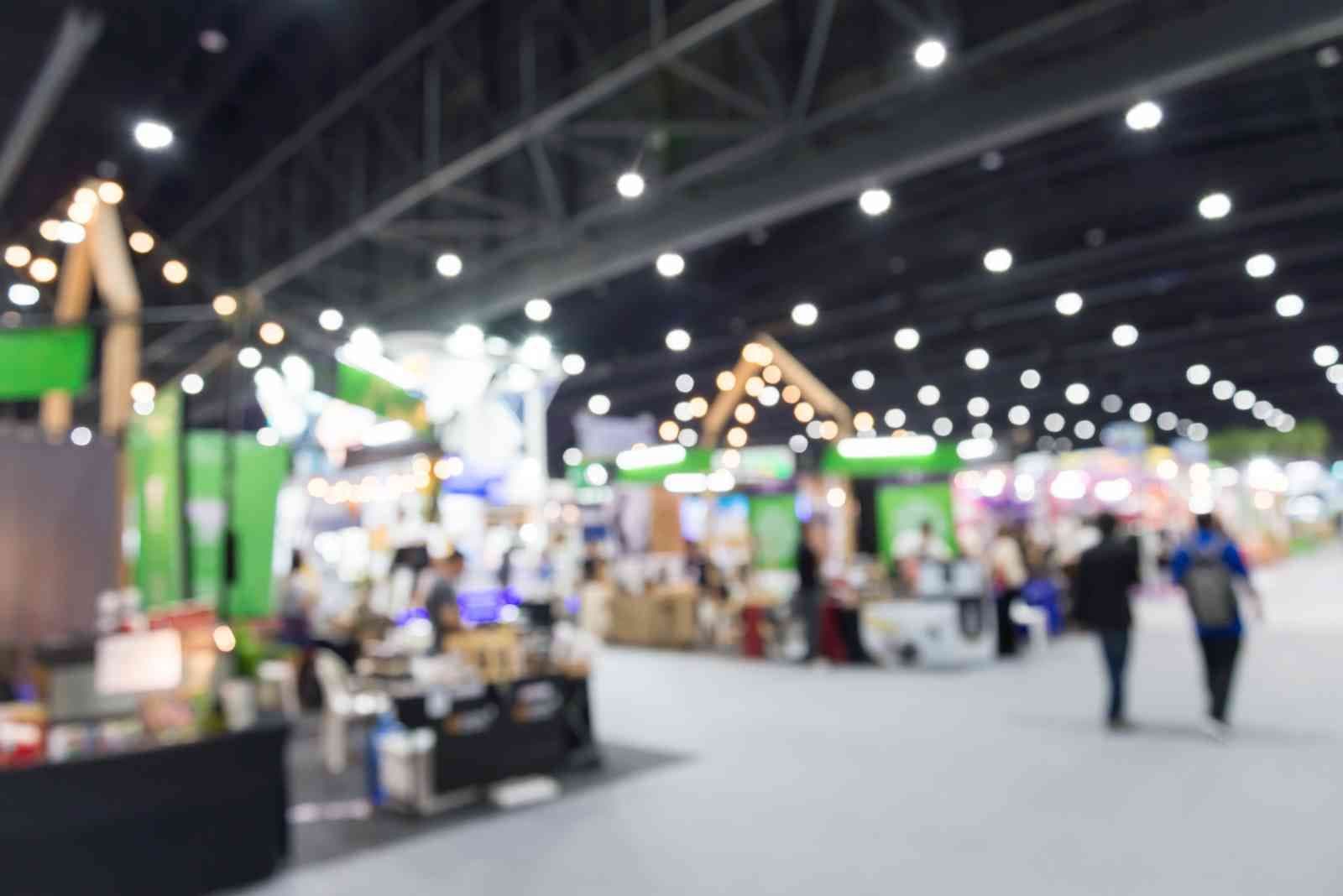 Blurred image of a bustling trade show with bright lights, colorful booths, and people walking around, creating a vibrant and busy atmosphere.