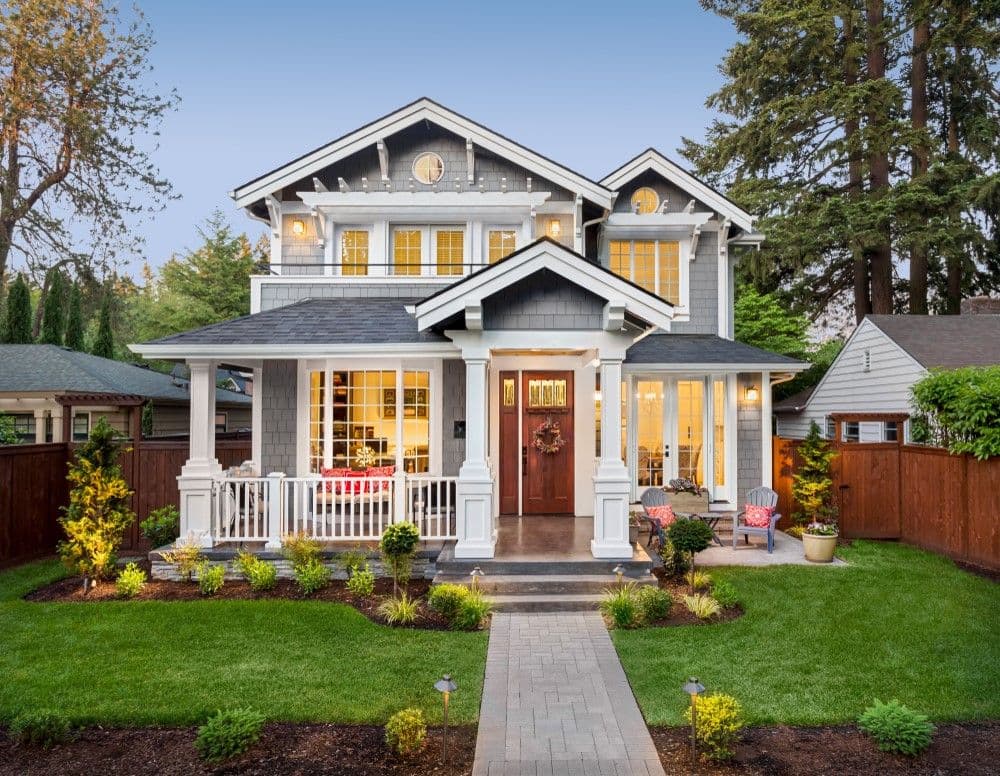 Charming two-story gray house with white trim, a front porch, manicured lawn, and warm lights glowing inside. A welcoming, serene suburban setting.