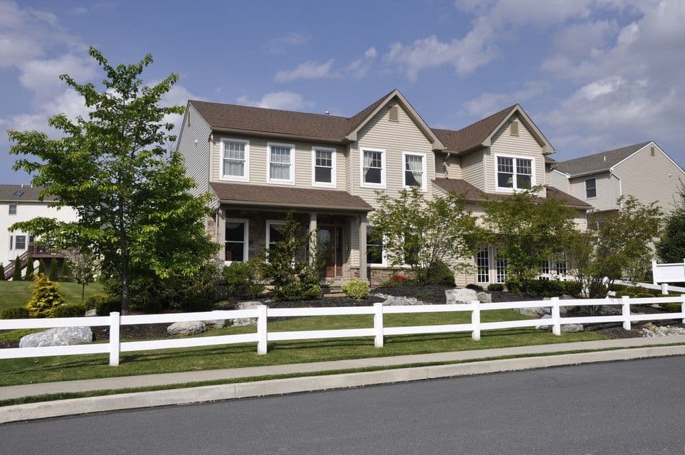 A spacious two-story beige house with a brown roof, surrounded by a neatly landscaped garden and white fence, under a partly cloudy blue sky.