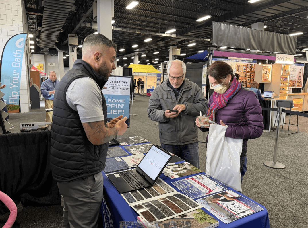 A vendor at a trade show booth discusses products with two attendees. The table displays samples and brochures. The atmosphere is busy and engaging.