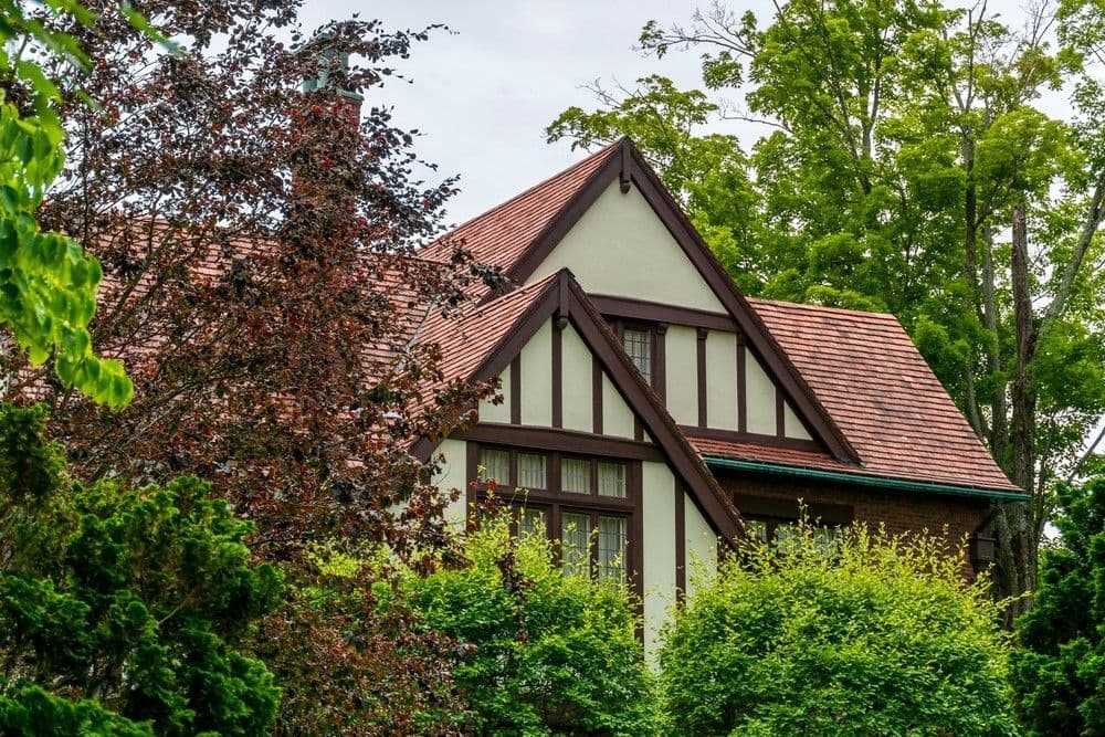 Tudor-style house with decorative timber framing and a steep red-tiled roof, surrounded by lush green trees and shrubs, evoking a serene atmosphere.