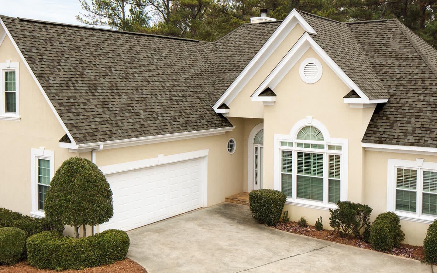 A beige suburban house with a dark shingle roof, white garage door, and arched window. The driveway is bordered by neatly trimmed bushes, evoking a tidy and welcoming feel.