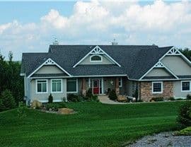 Single-story house with gray siding and stone accents, dark roof, and red front door. It is surrounded by a manicured green lawn under a partly cloudy sky.