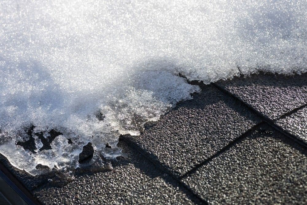 Close-up of melting snow on a dark shingle roof. The sunlit snow contrasts with textured shingles, creating a transient, wintry atmosphere.