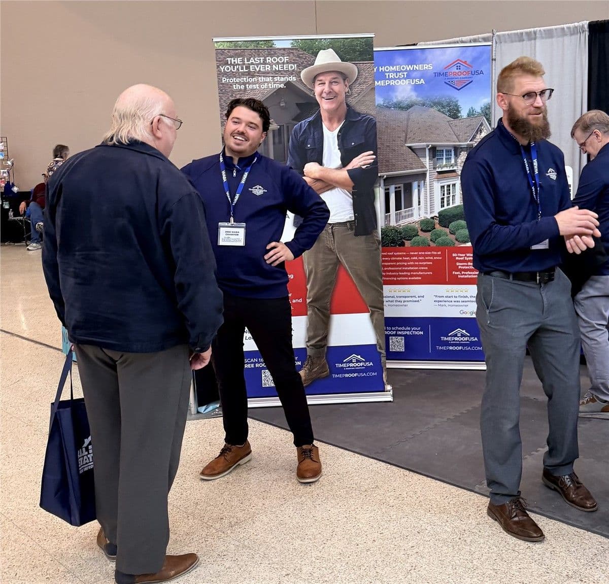 Three men in navy shirts converse at a tradeshow booth with roofing banners. They appear engaged and friendly, creating a welcoming atmosphere.