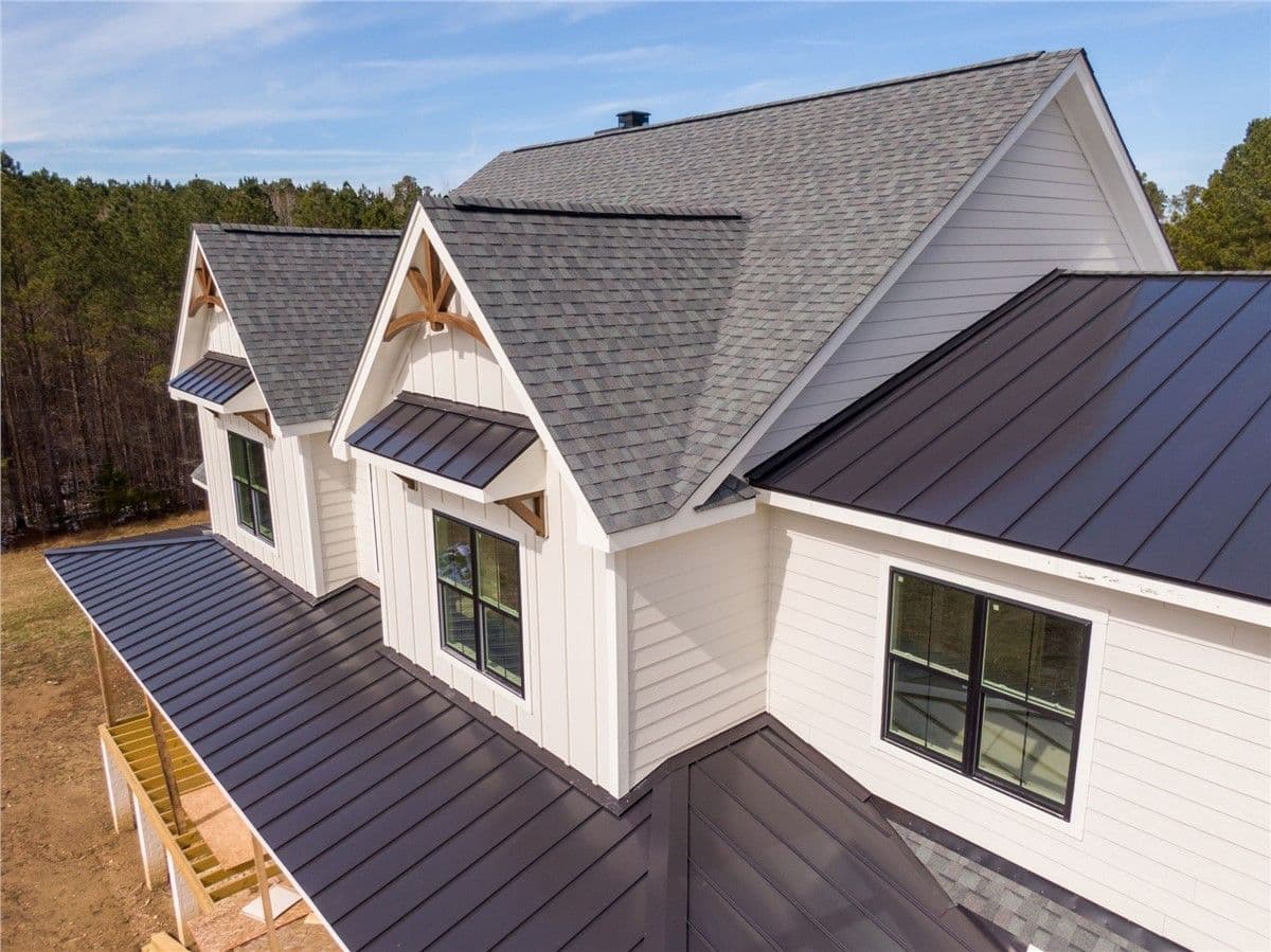 Aerial view of a modern house with a gabled roof, gray shingles, and white siding. Large windows and metal accents add to the contemporary design. Forest in the background.