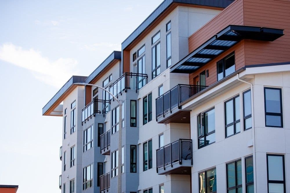 Modern multi-story apartment building with large windows and balconies, featuring a mix of white and wood paneling under a clear blue sky.