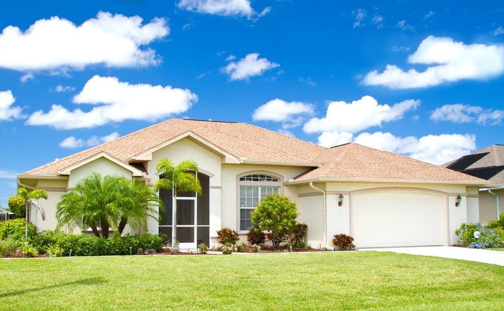 Single-story house with white walls, tan shingled roof, and arched windows, surrounded by vibrant green lawn and tropical plants under a bright blue sky.