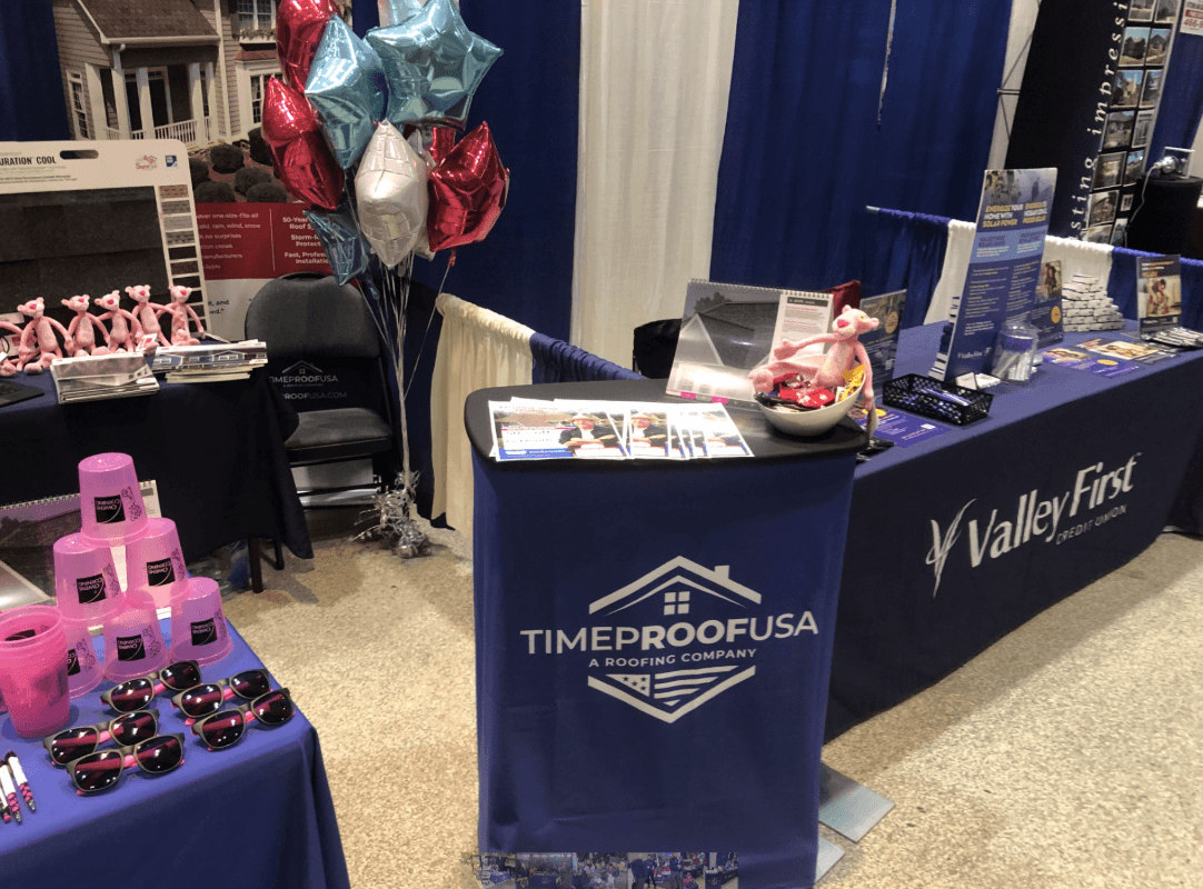 Exhibit booth with blue tablecloths featuring Time Proof USA and Valley First logos. Pink cups, sunglasses, and balloons create a festive, promotional mood.