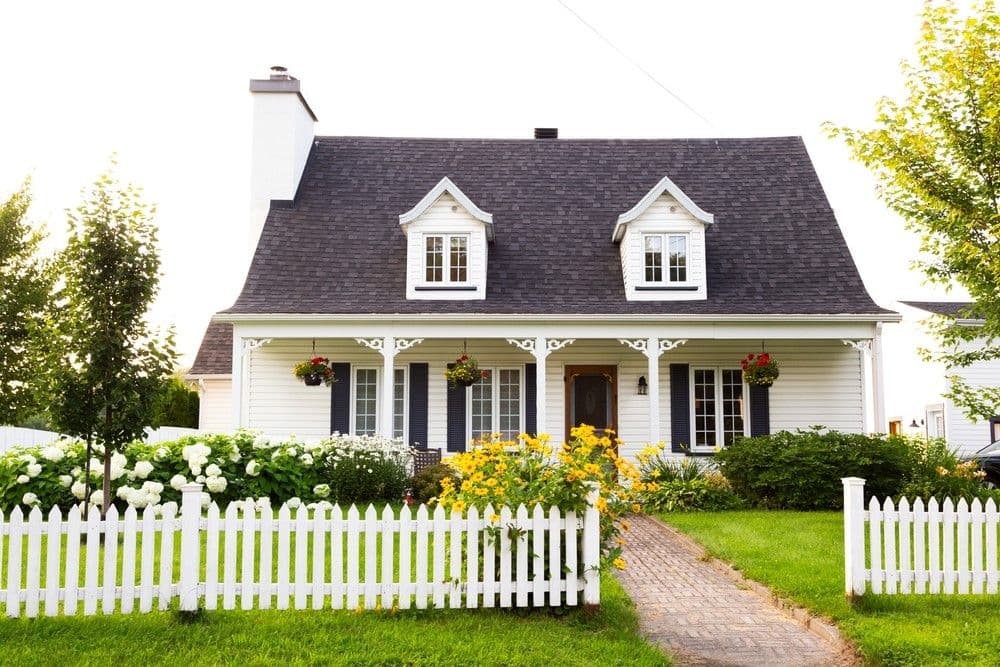 Charming white cottage with a dark roof, dormer windows, and a front garden lush with flowers. White picket fence and a brick path add to its cozy feel.