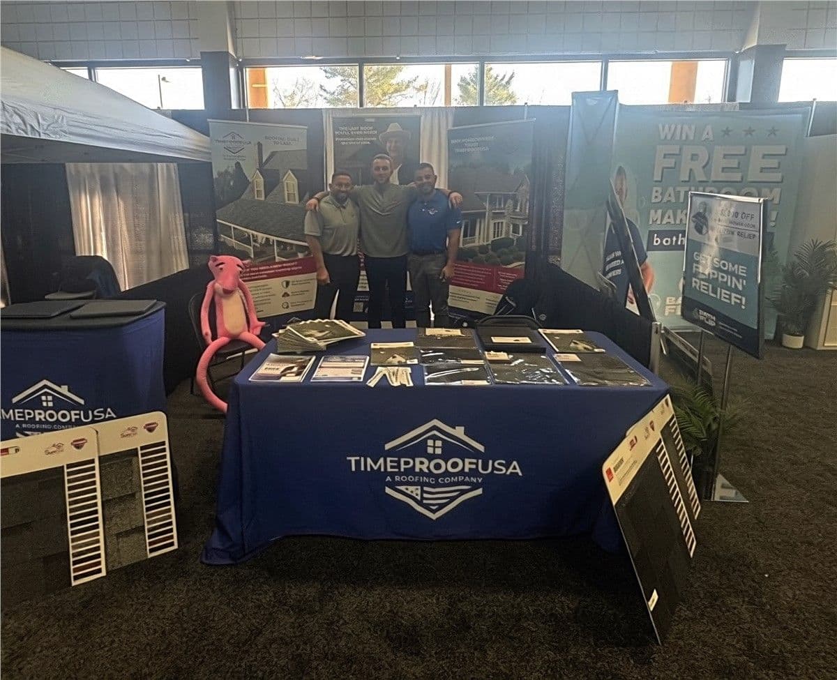Three people stand smiling behind a booth with "TimeProofUSA" branding. The booth displays roofing samples and brochures. A pink panther figure sits nearby. The atmosphere is professional and welcoming.