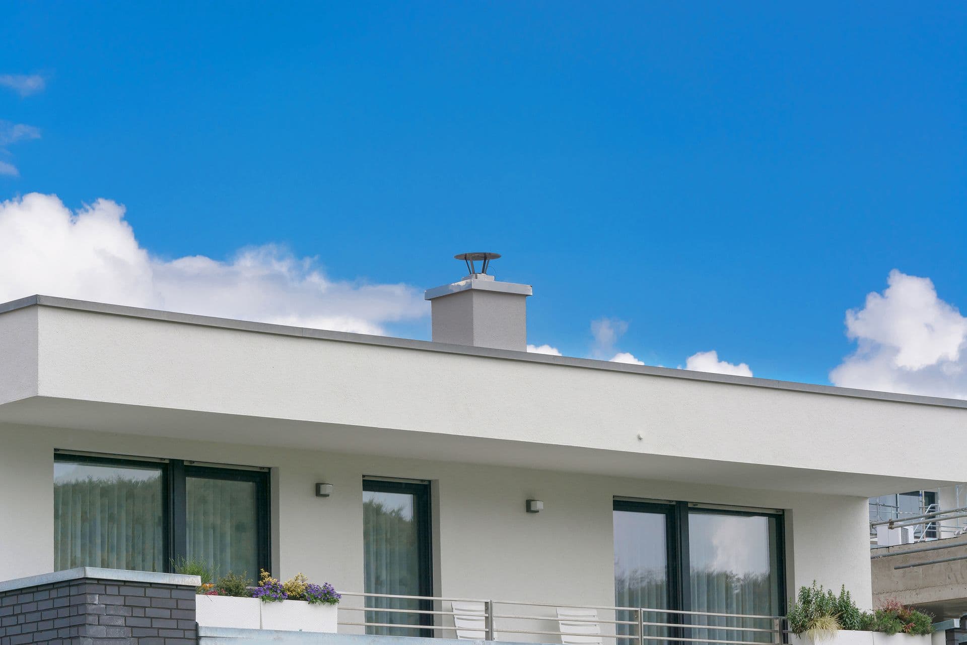 Modern white building with large windows, a flat roof, and a chimney under a clear blue sky. Flower boxes add a touch of color, conveying tranquility.