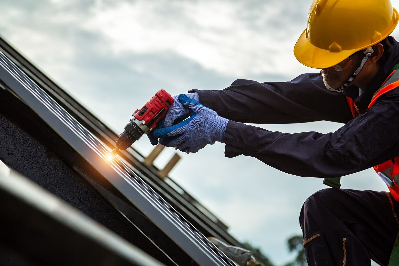 A construction worker in a hard hat and safety vest uses a power drill on a rooftop. The image conveys focus and precision in a work setting.