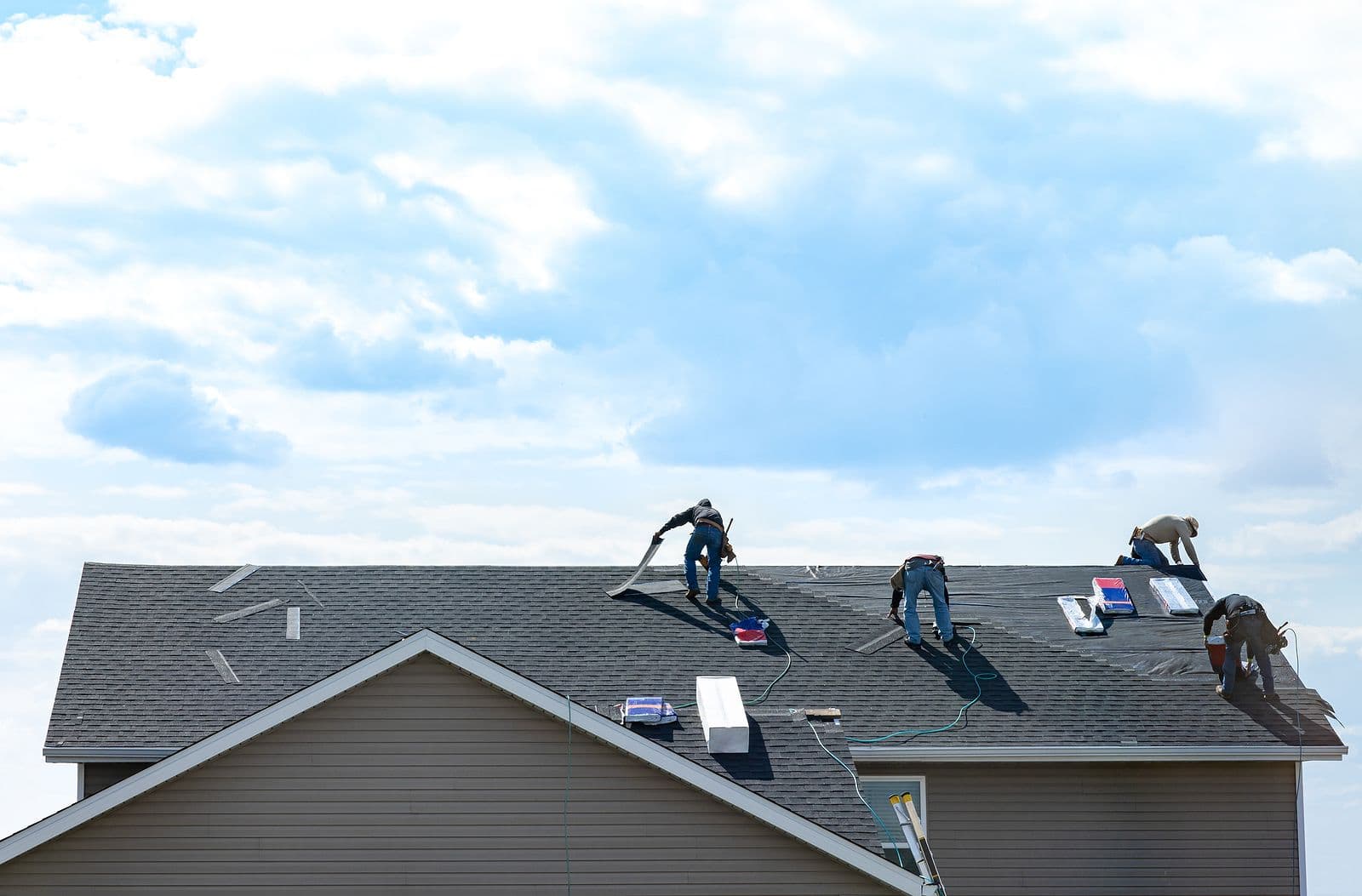 Workers repair shingles on a rooftop against a bright blue sky with clouds. The scene conveys industriousness and teamwork under sunny conditions.