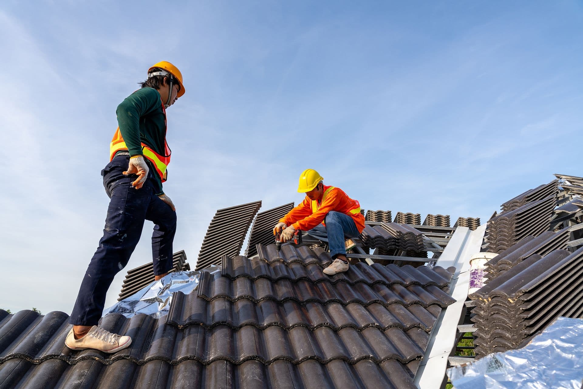 Two construction workers wearing hard hats and safety vests are installing roofing tiles on a house under a clear blue sky, conveying teamwork and focus.