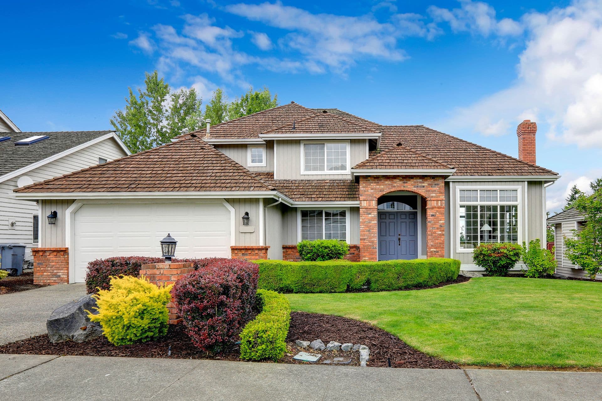 Two-story suburban house with brick accents and a blue front door. Manicured lawn and colorful shrubs frame the entrance under a clear blue sky.