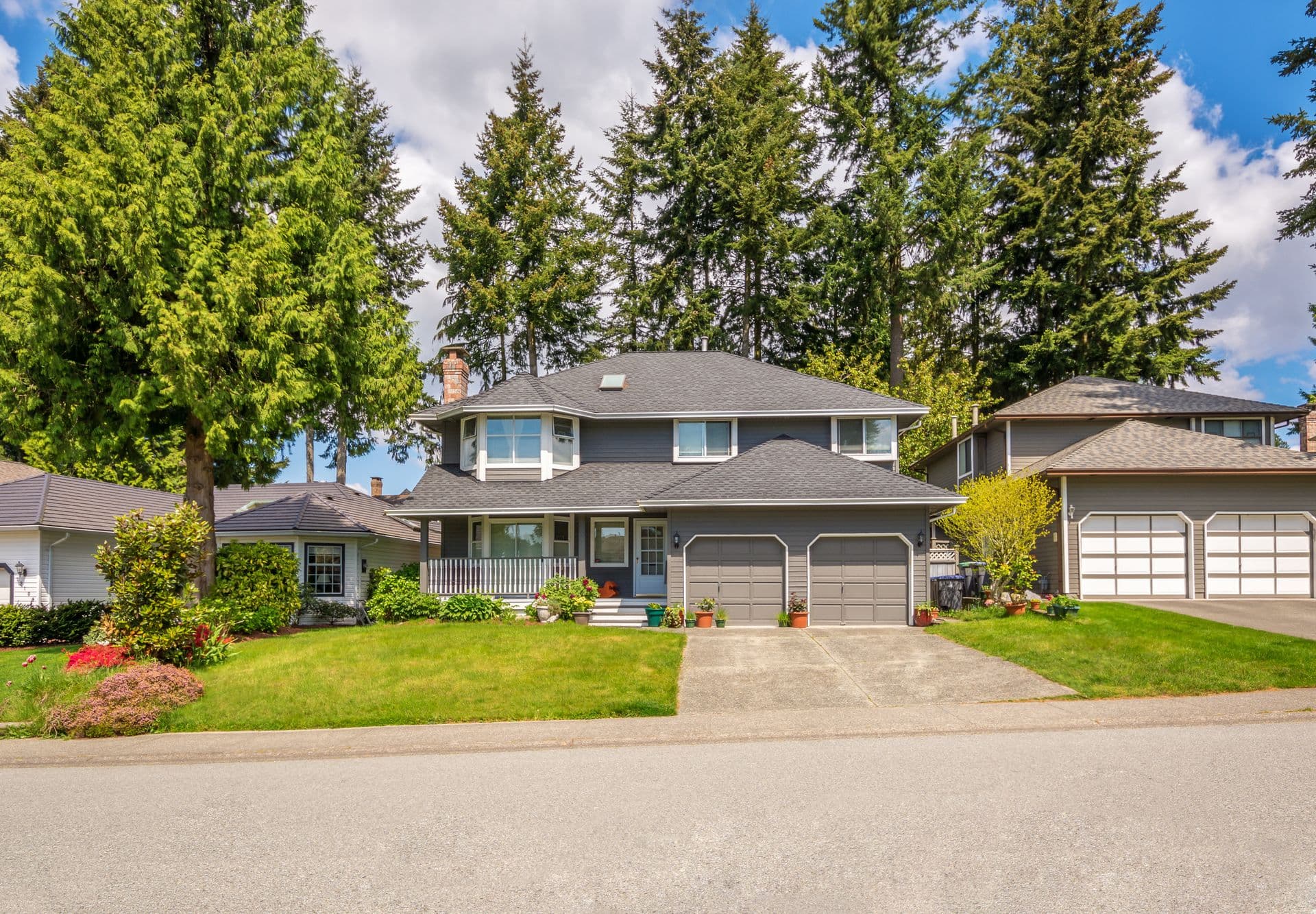 A two-story suburban house with gray siding, a neatly trimmed lawn, and vibrant flower pots. Surrounded by tall trees under a clear blue sky.