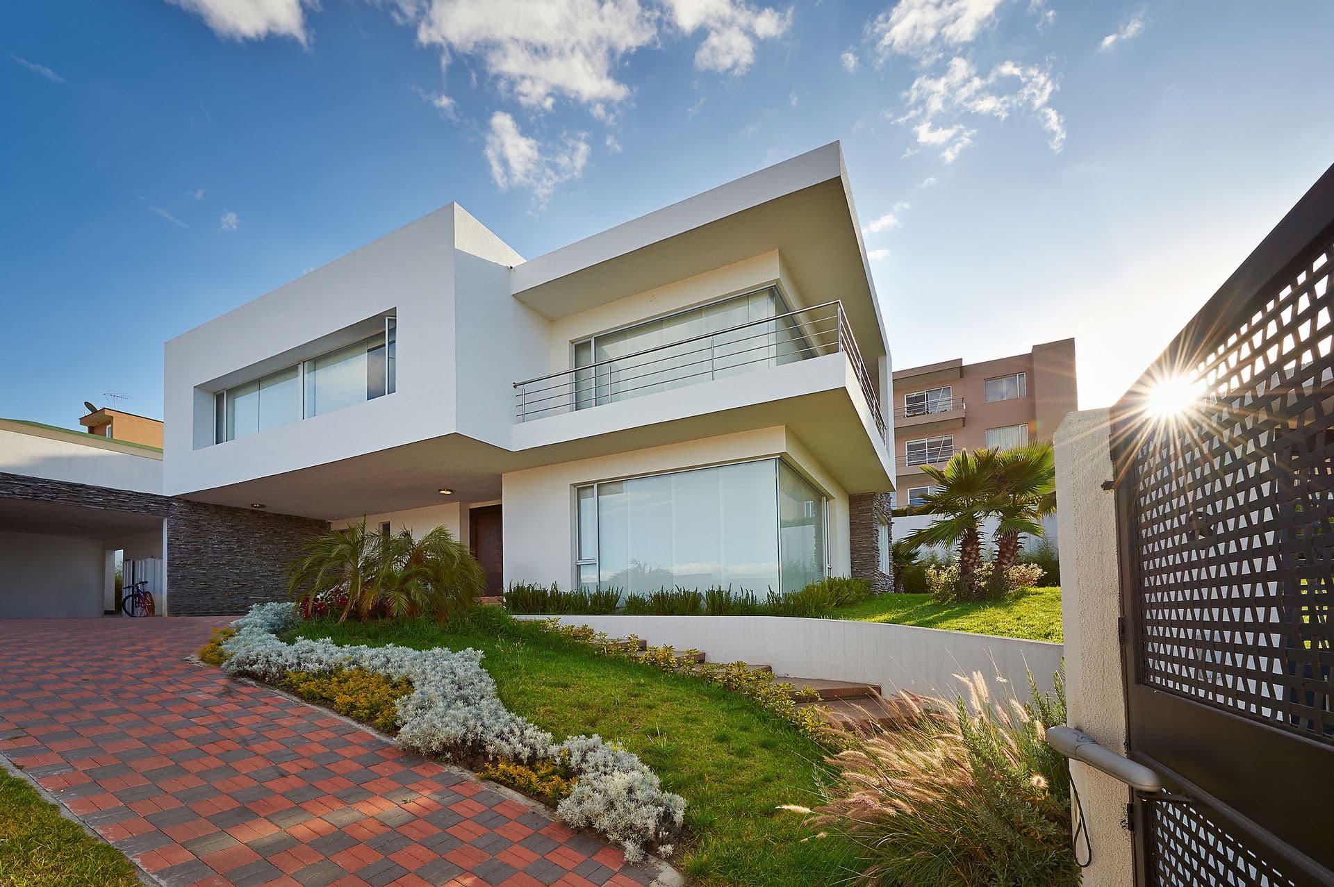 Modern two-story house with white walls and large windows. The front yard features lush greenery and a red-bricked path. Sunlight shines from behind.