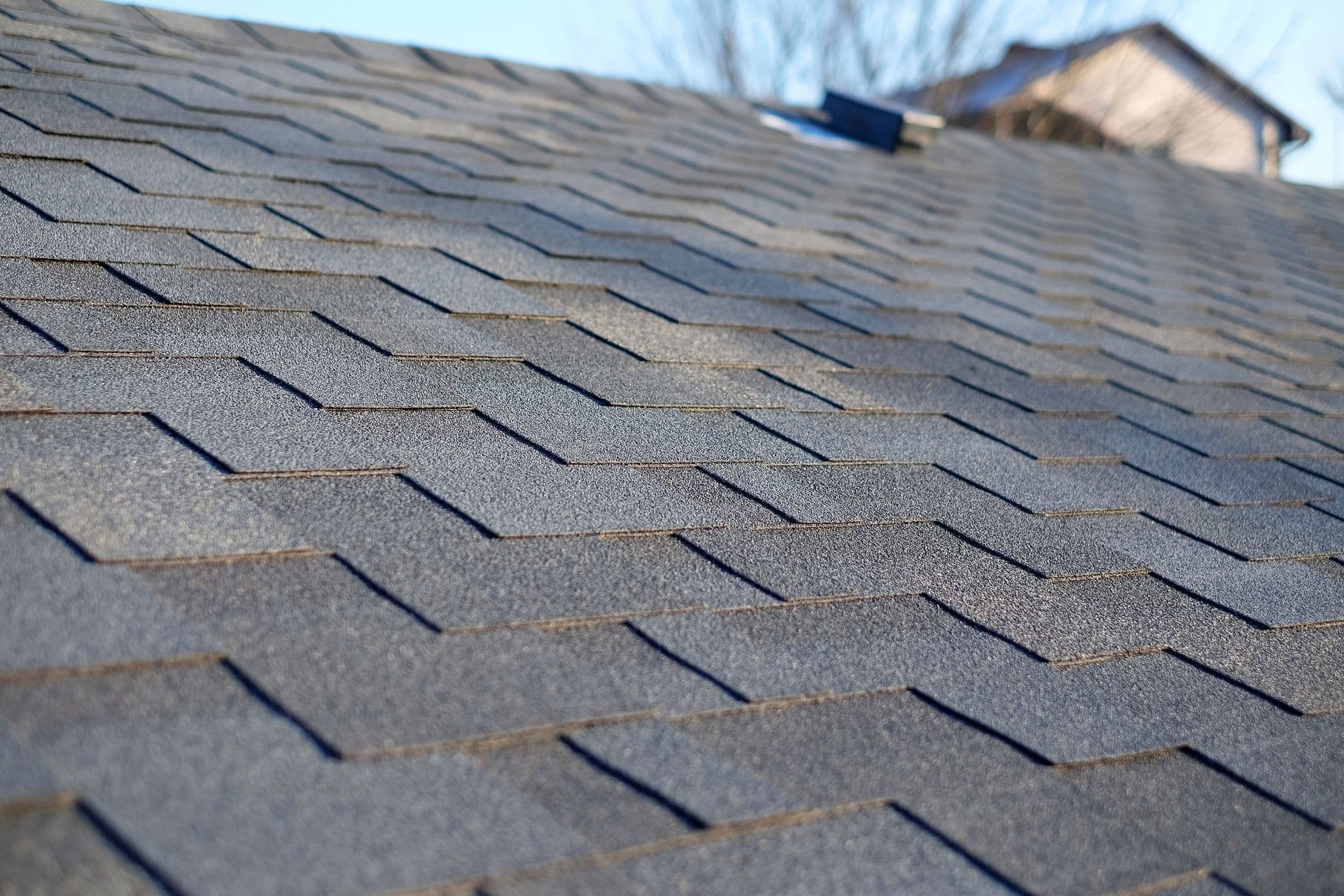 Close-up of a gray shingle roof in sunlight, showing the textured, overlapping pattern. In the background, part of a house roof is visible under a clear sky.