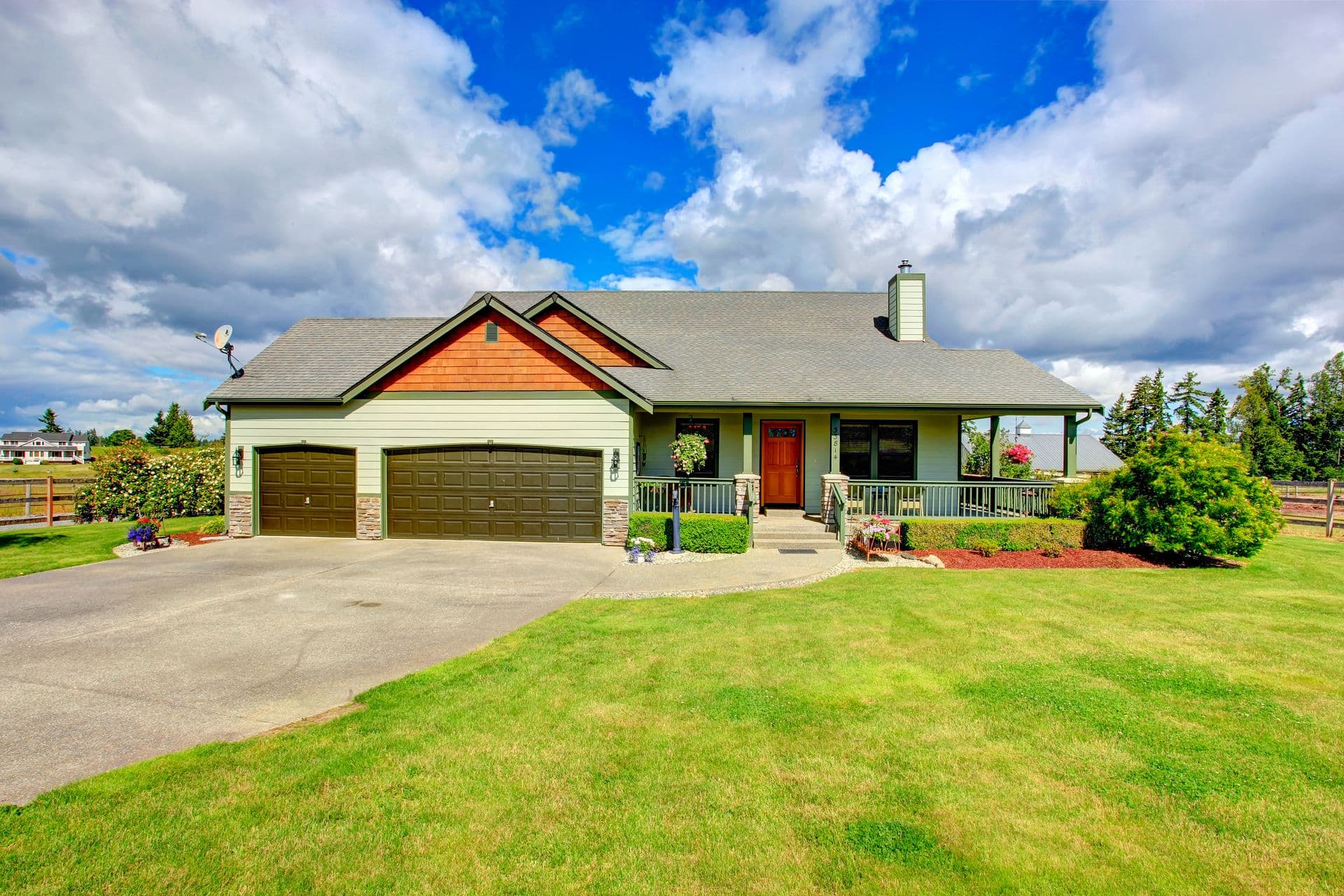 Charming suburban house with a gray roof, green siding, brown garage doors, and a red front door. Sunny day with a vivid blue sky and white clouds.