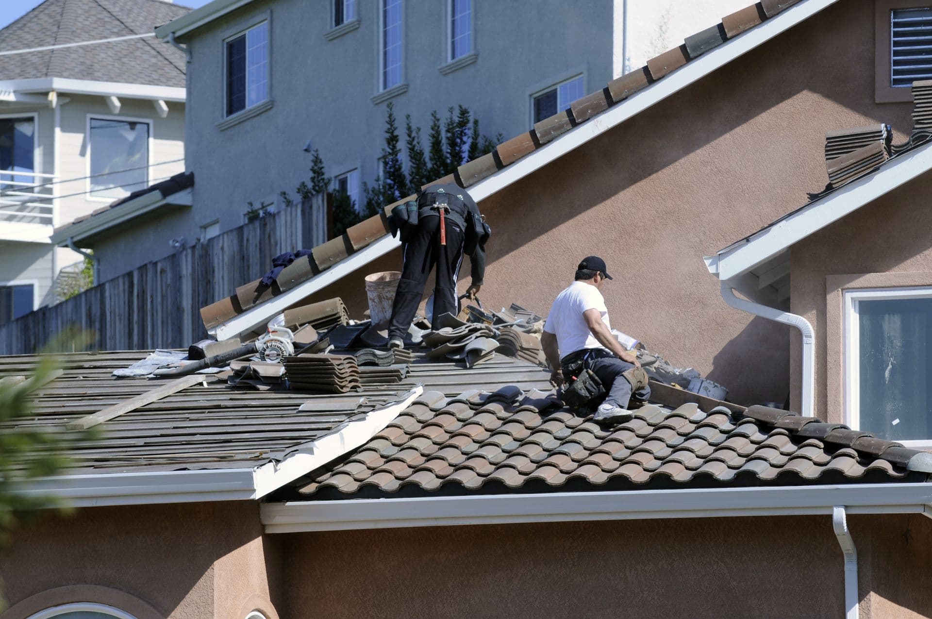 Two workers repair a tiled roof under clear skies, surrounded by tools and tiles. Nearby houses hint at a suburban setting. The mood is industrious.