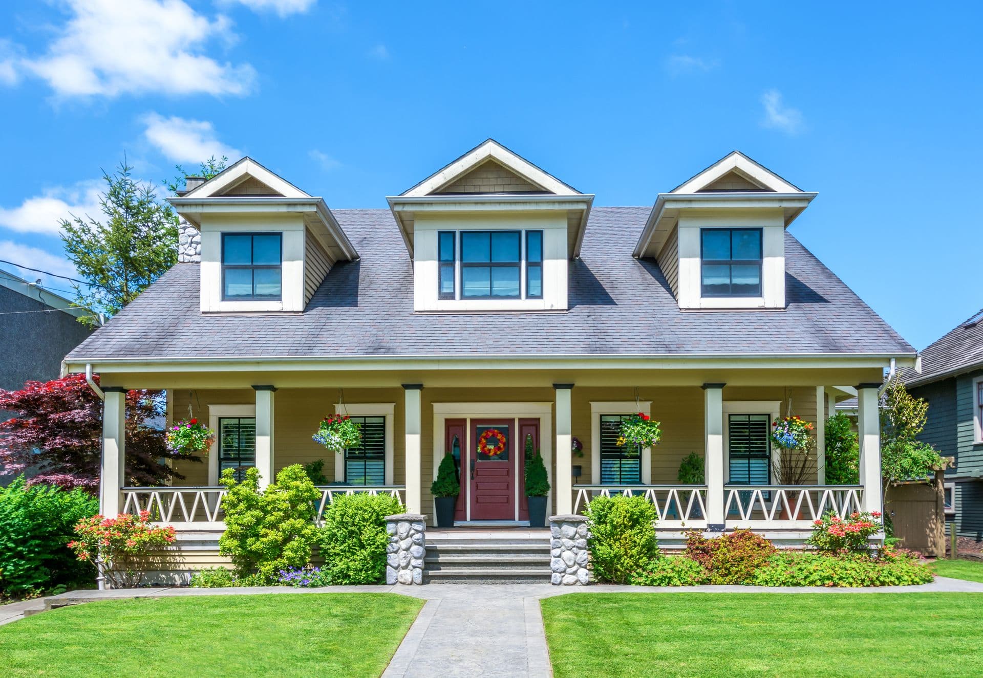 A charming yellow house with a dark roof, featuring three dormer windows and a wide porch. Surrounded by green lawn, shrubs, and colorful flowers.