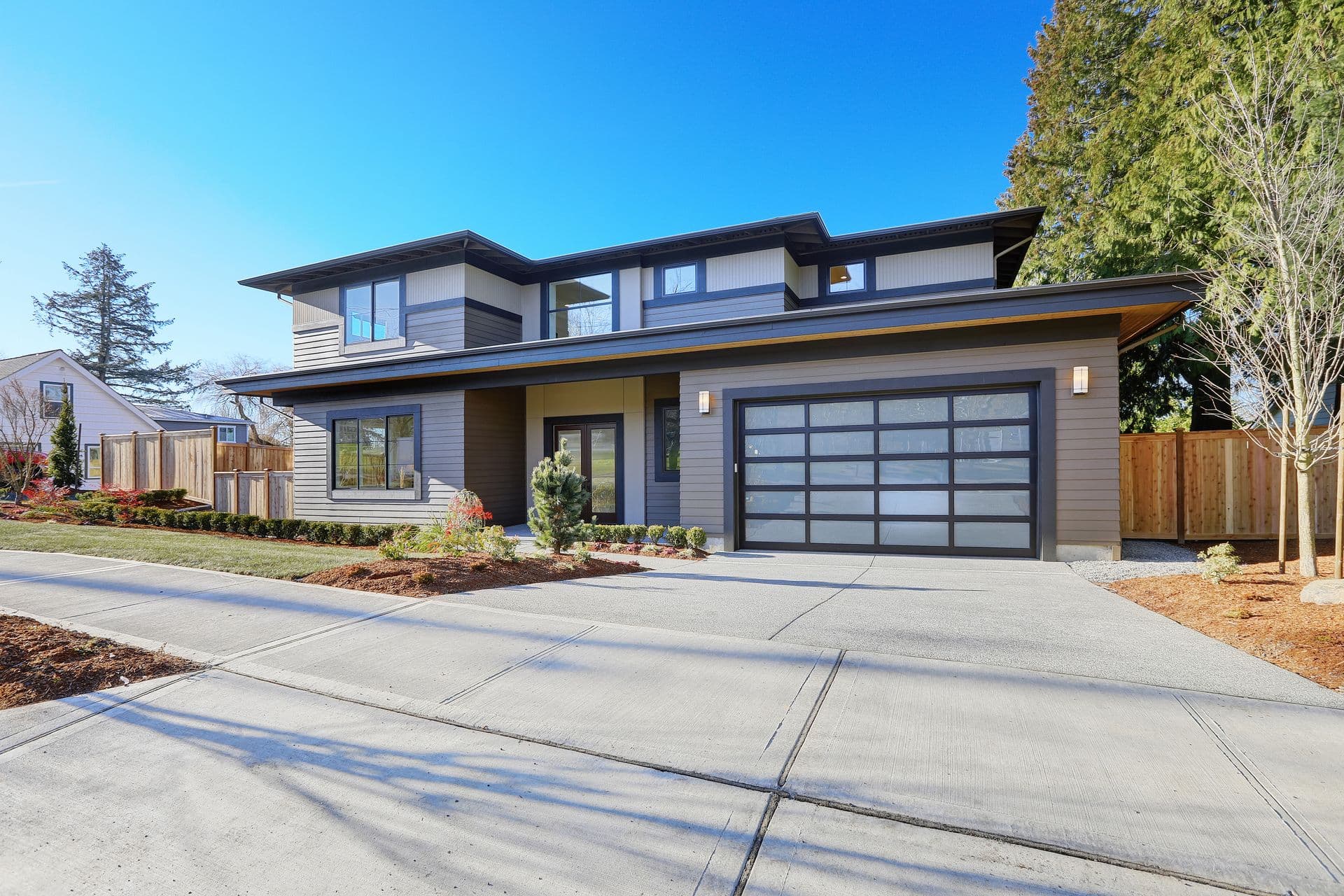Modern two-story house with a gray facade, large glass garage door, and landscaped front yard under a clear blue sky, conveying a sleek, contemporary feel.