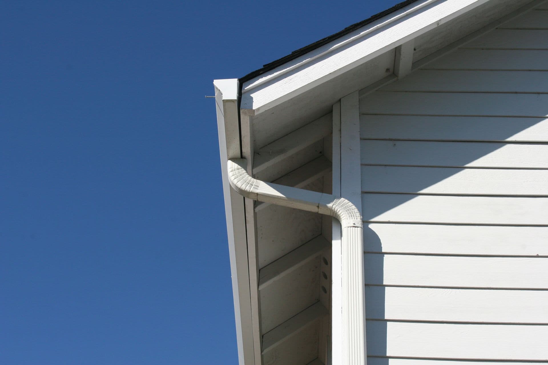 A white house exterior with a sloped roof and attached white gutter under a clear blue sky, creating a clean, minimalist architectural look.