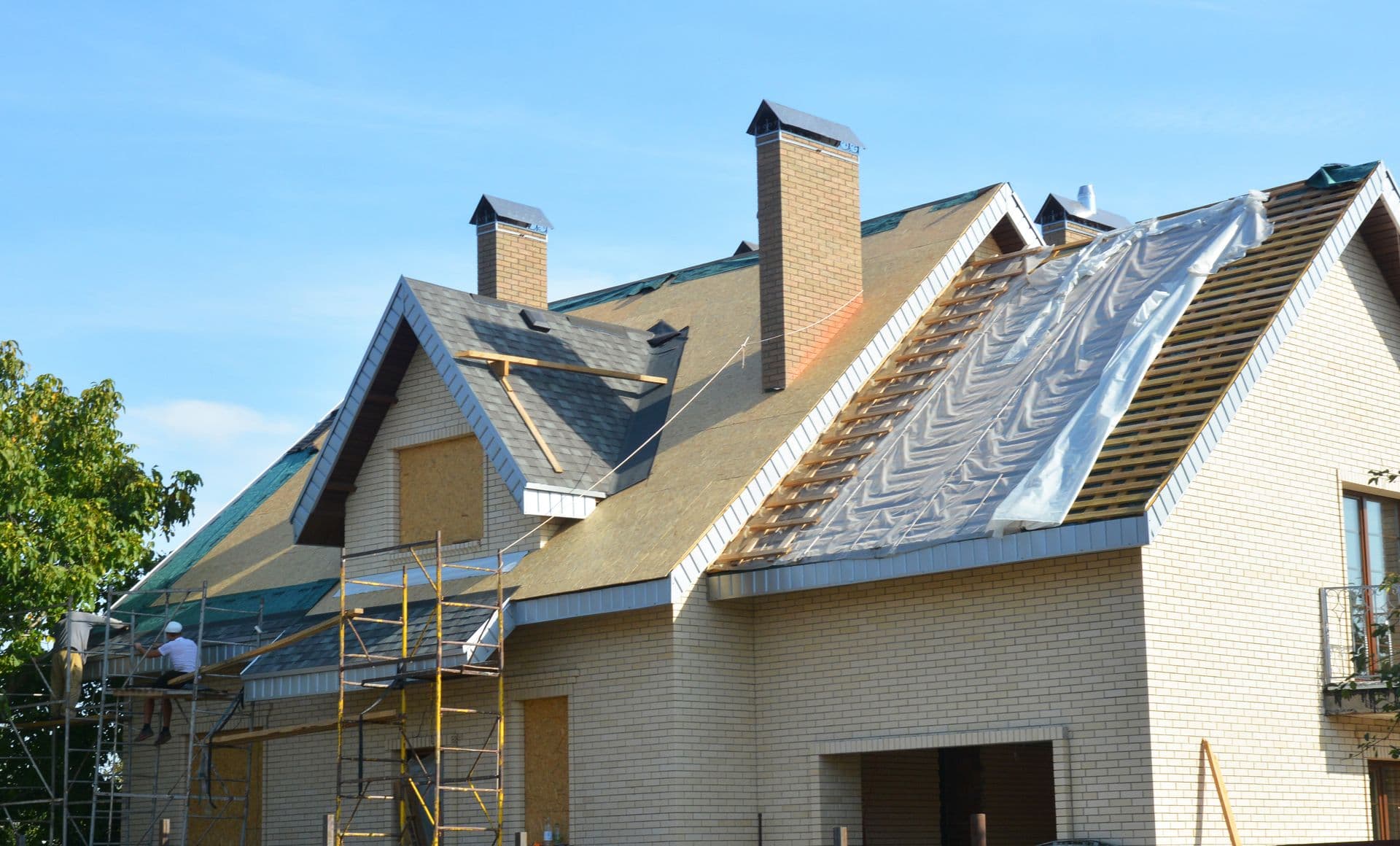 A brick house with scaffolding, under construction. Workers repair a steep roof, partially covered with a tarp. Clear blue sky in the background.