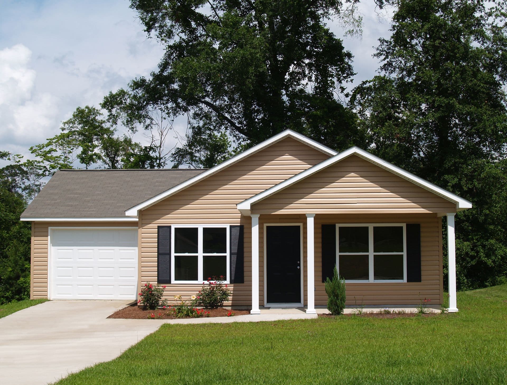 Single-story beige house with white trim, black shutters, and a dark front door. Attached garage, surrounded by green lawn and tall trees. Bright, serene atmosphere.