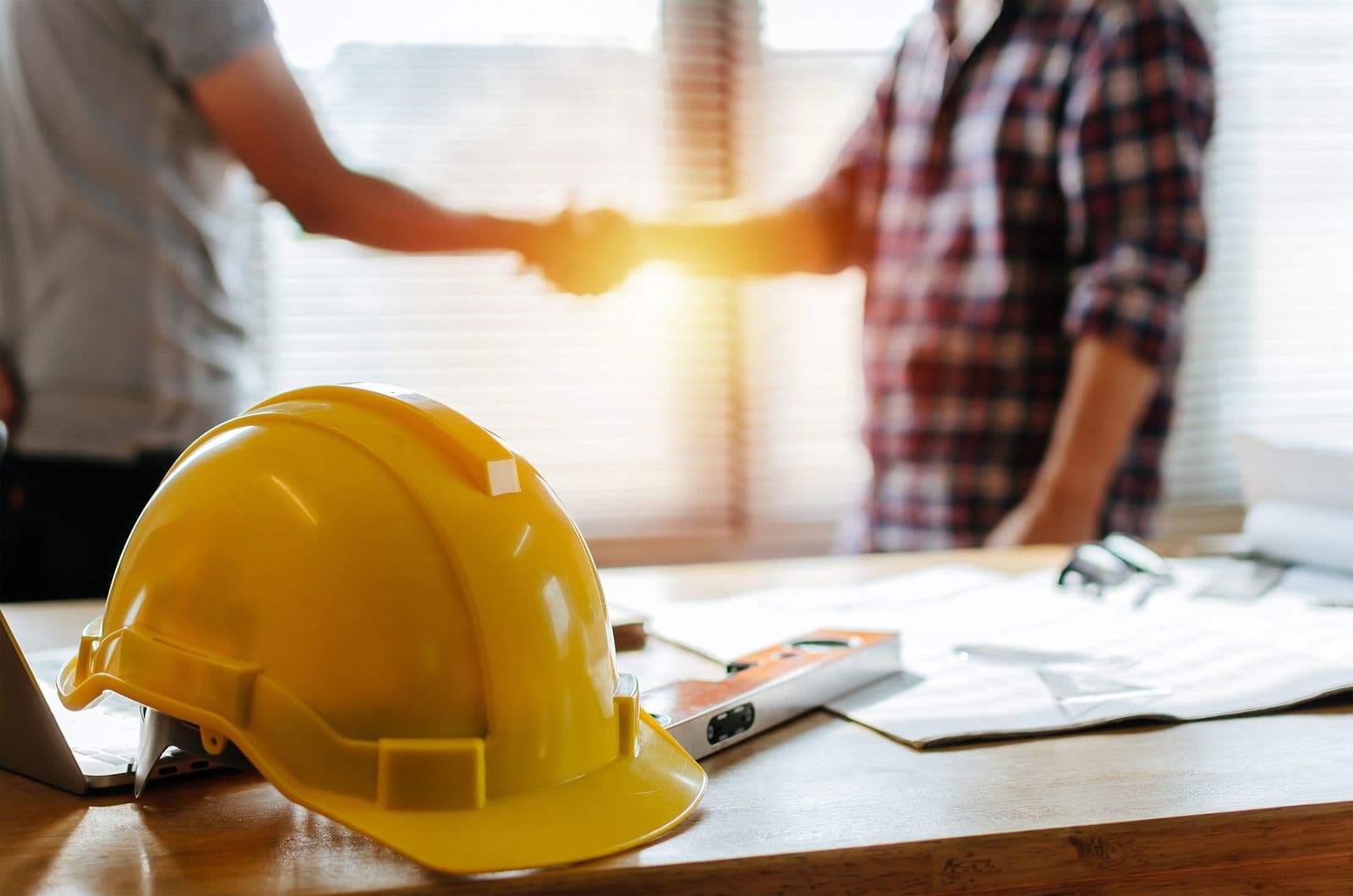 Yellow hard hat on a desk with construction plans; two people shake hands in the background, symbolizing agreement. Sunlight filters through blinds.