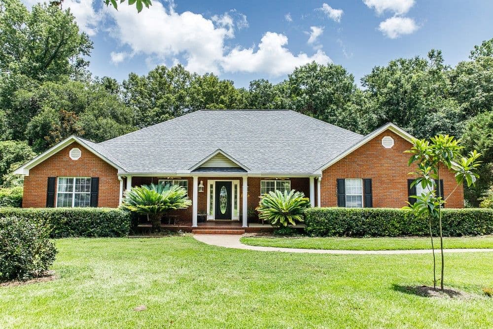 A charming brick house with a gray roof sits amidst a lush green lawn, framed by symmetrical bushes. The sky is blue with fluffy clouds above.