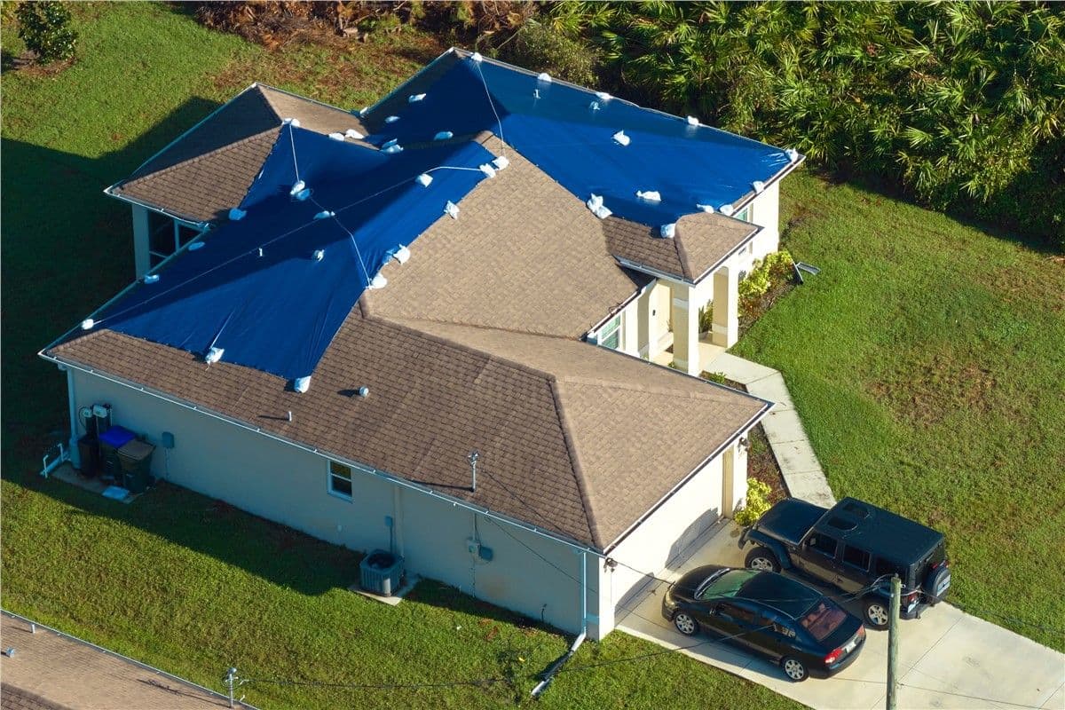 Aerial view of a house with multiple triangular blue tarps covering sections of the tan roof. Two cars are parked in the driveway, surrounded by green grass.