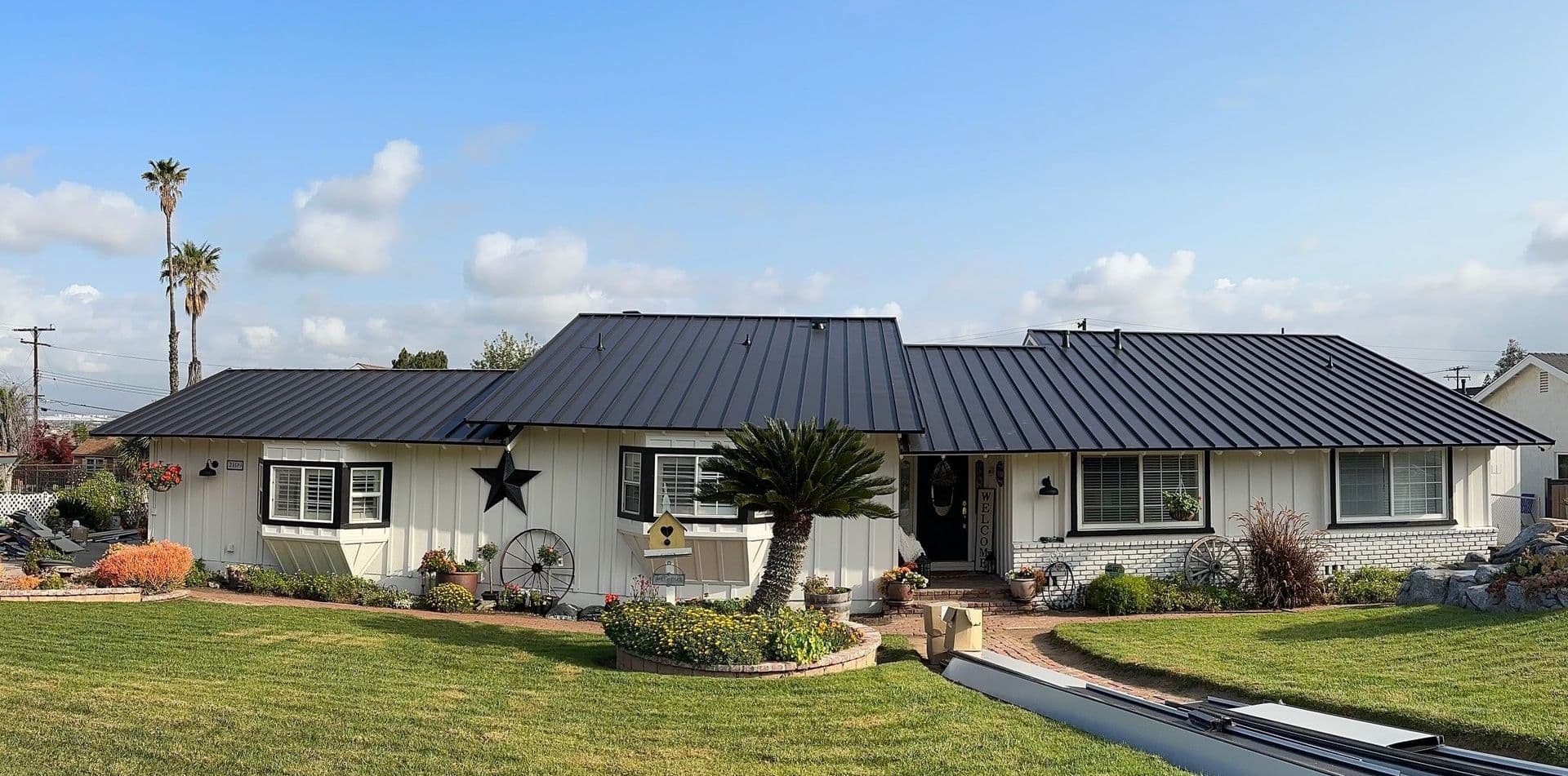 One-story house with a black metal roof, white siding, and large windows. The front yard features neatly trimmed grass, plants, and a birdhouse. The sky is partly cloudy.