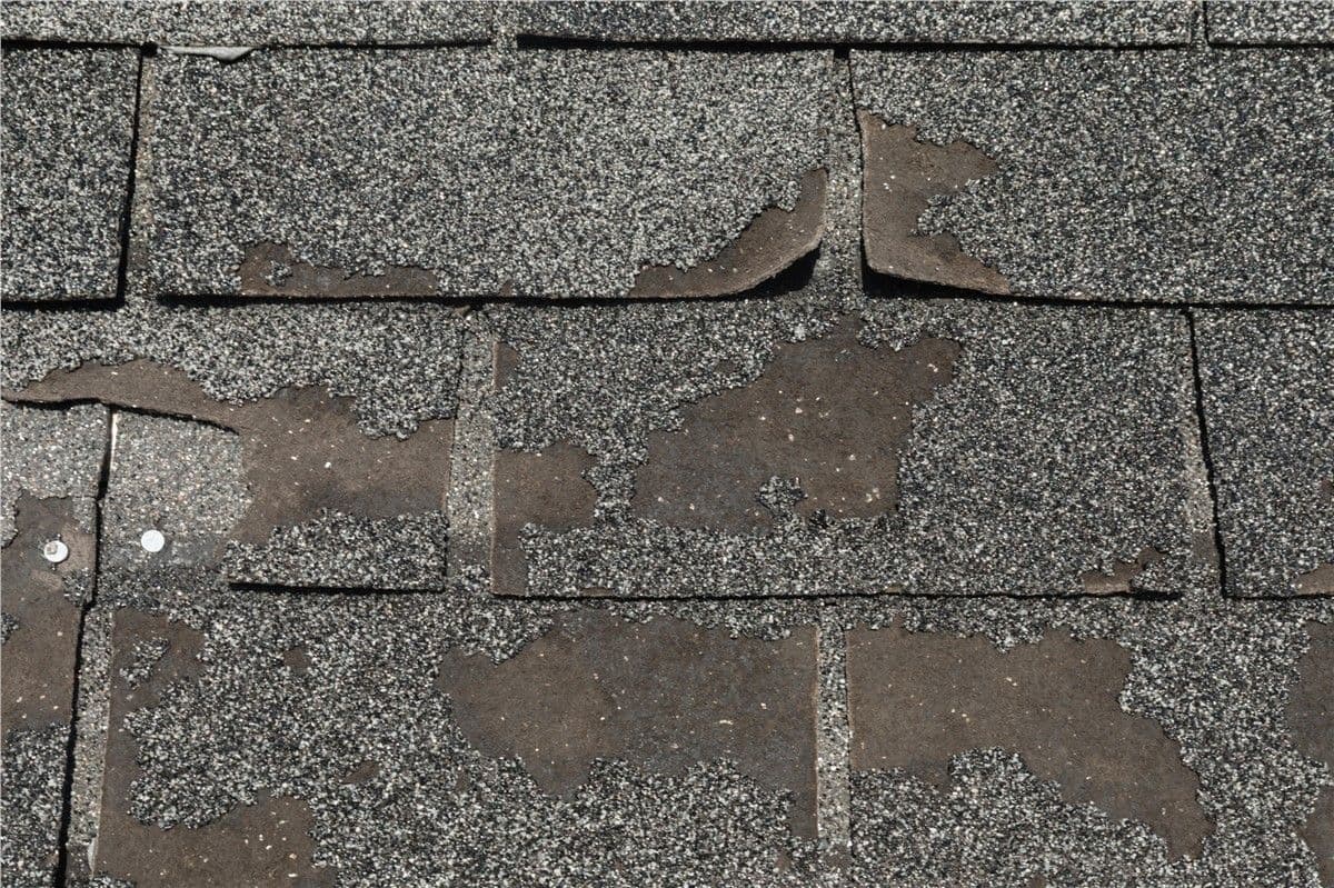 Worn asphalt shingles with peeling edges and missing granules, revealing dark underlayer. Conveying age, neglect, and need for roof repair.