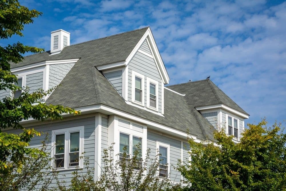 A charming two-story house with gray siding and white trim against a partly cloudy blue sky. Lush green trees frame the scene, conveying a peaceful suburban feel.