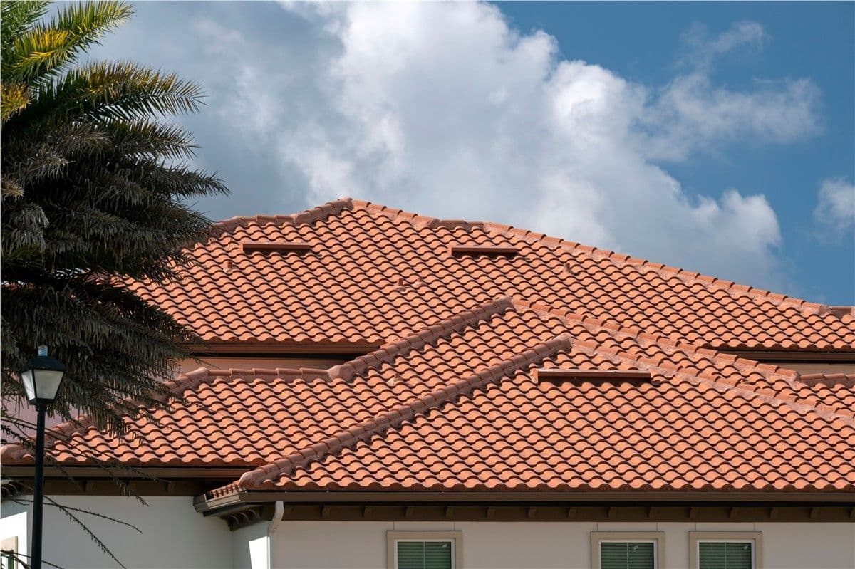 Red clay tile roof of a house under a blue sky with fluffy clouds. A streetlamp and palm tree are partially visible on the left, conveying a warm, tranquil atmosphere.