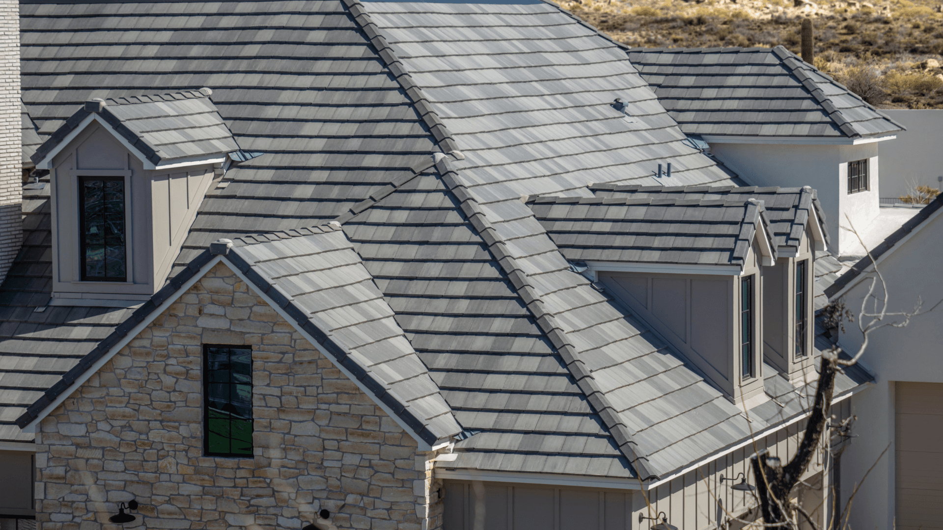 Steeply-sloped gray slate metal roof on a house with dormer windows and stone facade. The setting is bright and sunny, evoking a modern, sturdy design.