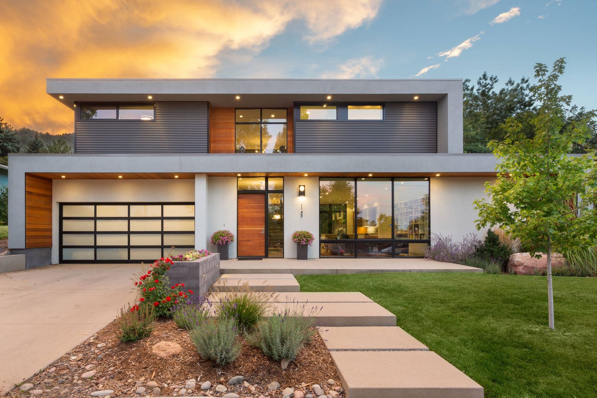 Modern two-story house with large windows and wood accents, surrounded by a manicured lawn and garden. Warm sunset sky enhances a serene ambiance.