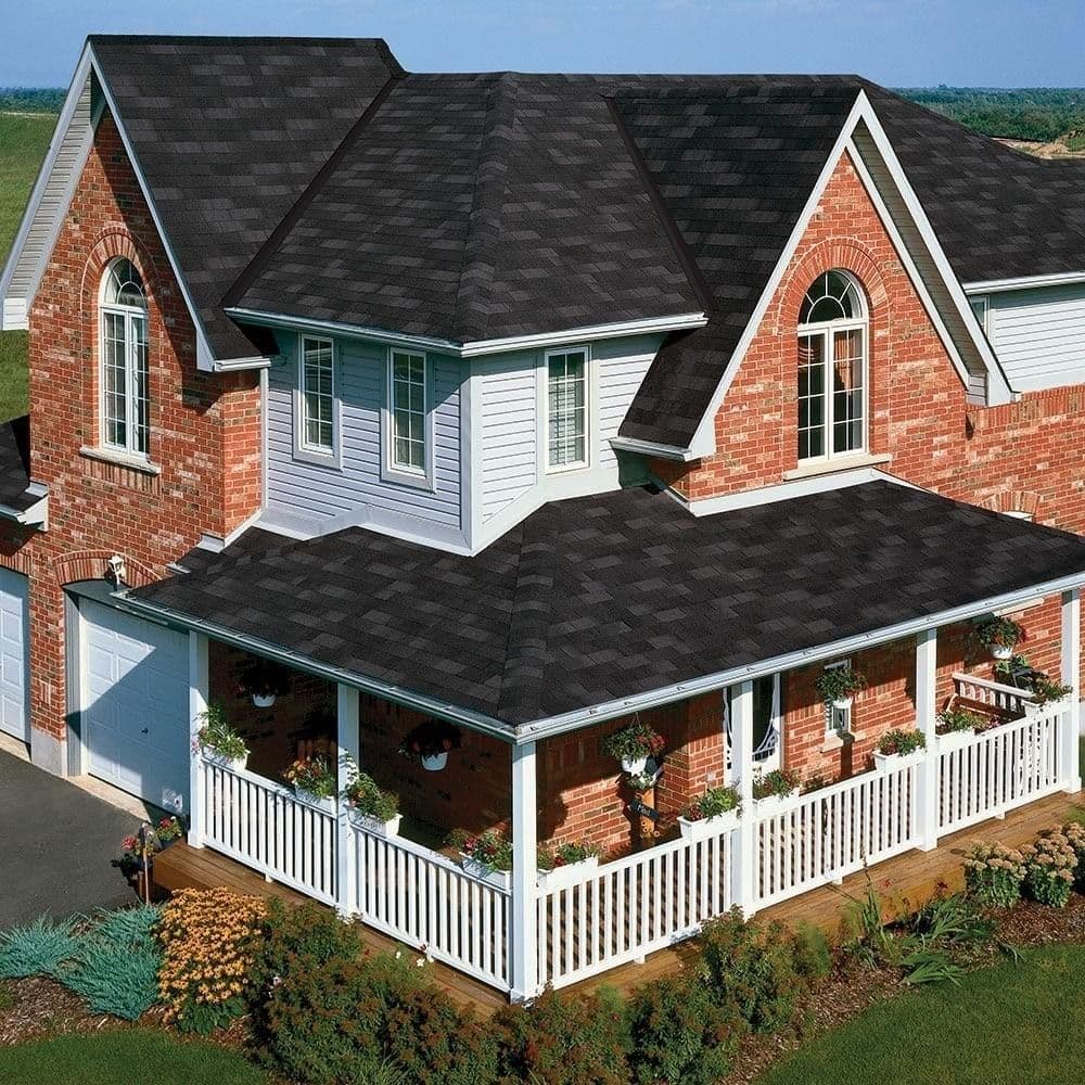 A charming two-story brick house with gray shingles, white trim, and an arched window. It features a wraparound porch with potted plants and a white fence.