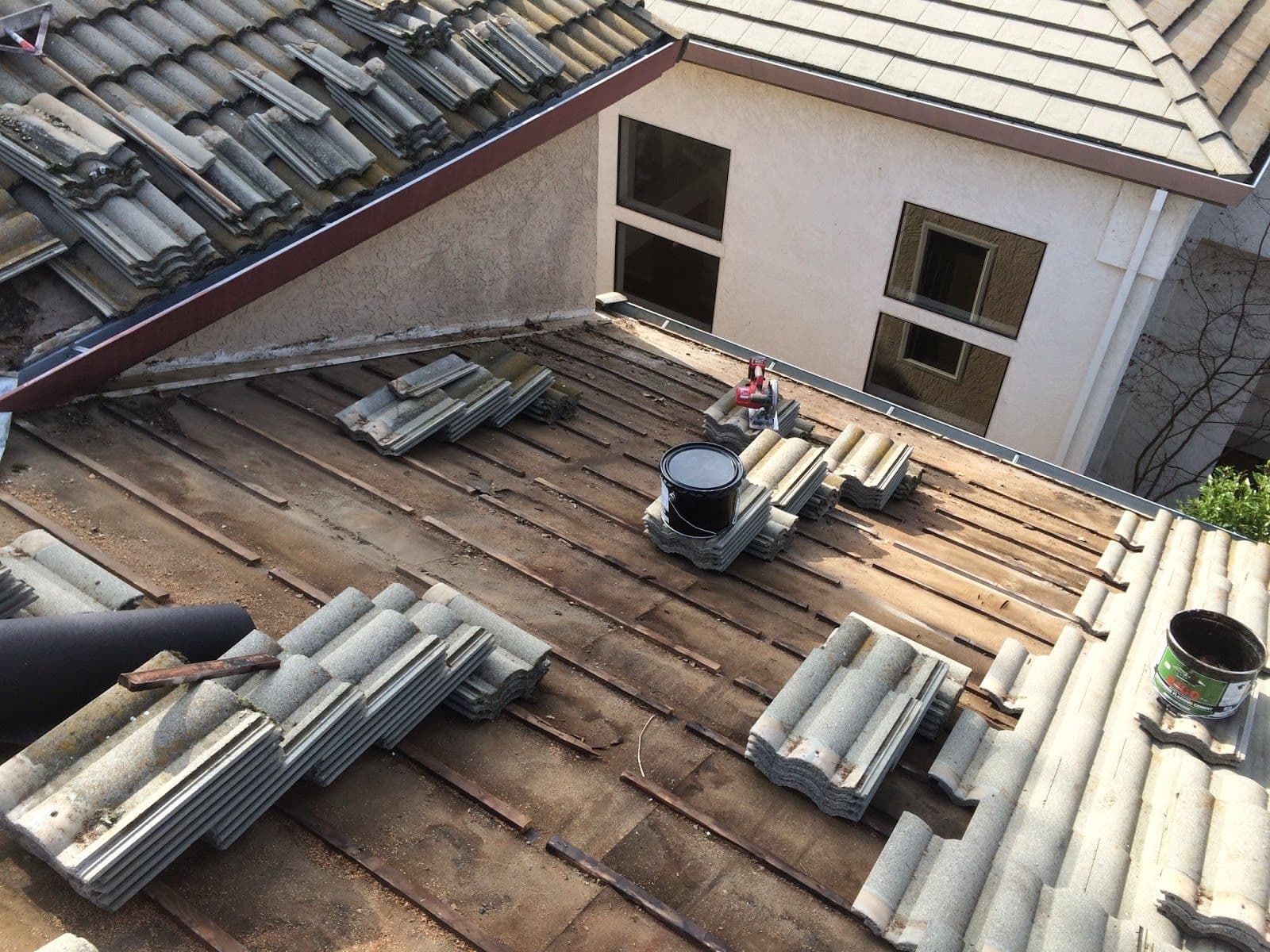 A partially dismantled roof with stacks of tiles and paint cans scattered around. Adjacent buildings have intact roofing. The area looks under renovation.